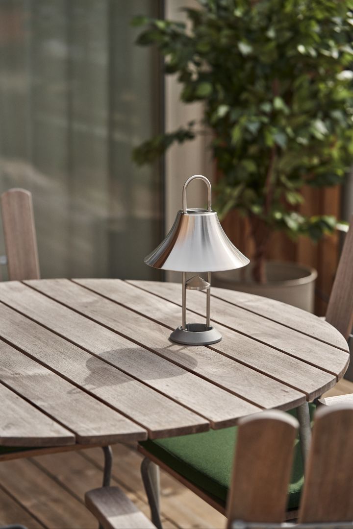 Få stemningsfullt lys til hagen eller terrassen med Mousqueton bærbar lampe i rustfritt stål fra HAY, som står på et rundt utebord i teak.