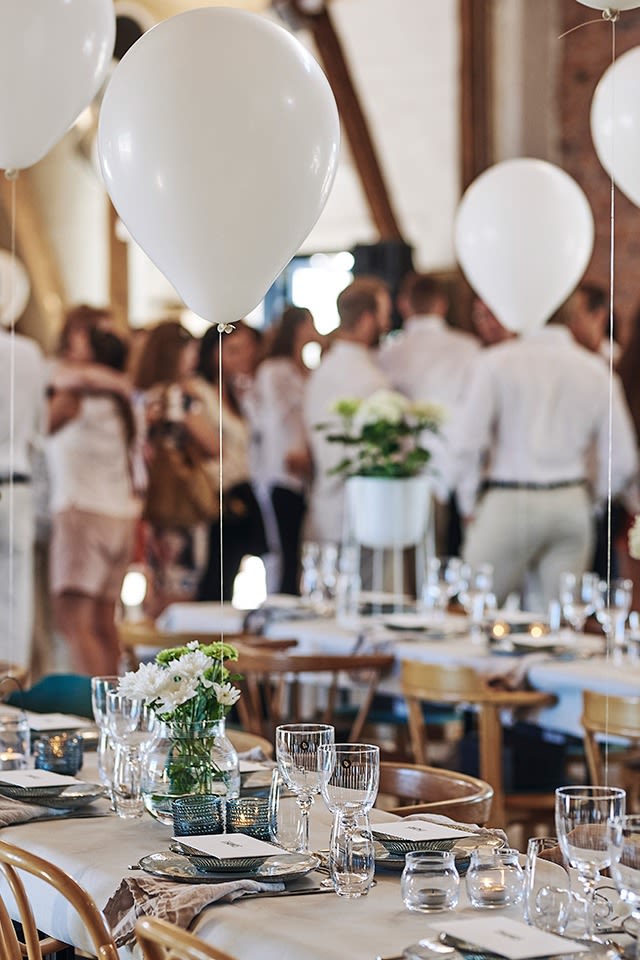 Langbord er satt opp til sommerfest i hvitt og bordet er dekket til mange mens ballonger svever over bordene. 