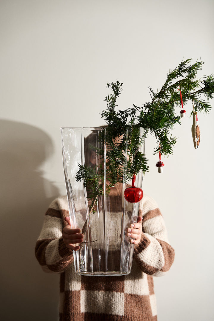 Hendene holder ut Reed-vasen fra Orrefors med vintage julepynt hengende fra en furugrein.